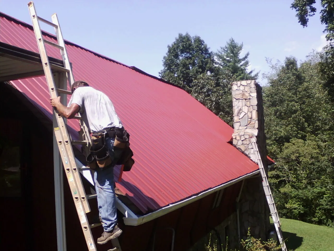 Roofer on a ladder beside a red metal roof and stone chimney — Soffit & Fascia Repair by Roofing Brothers in Boones Mill